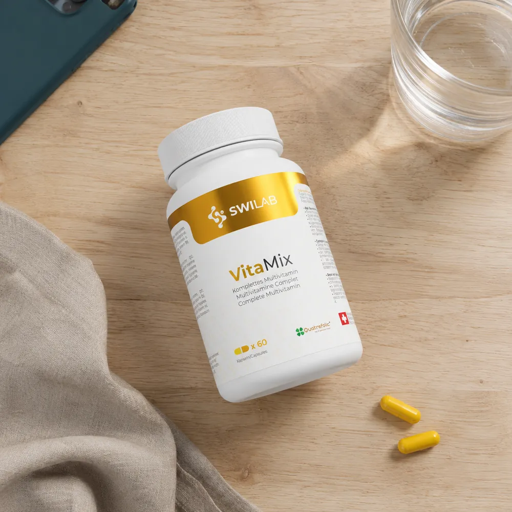 Flacon de multivitamines SwiLab vu de dessus sur une table en bois, entre un téléphone, un verre d'eau, un linge de lin et deux gélules jaunes.