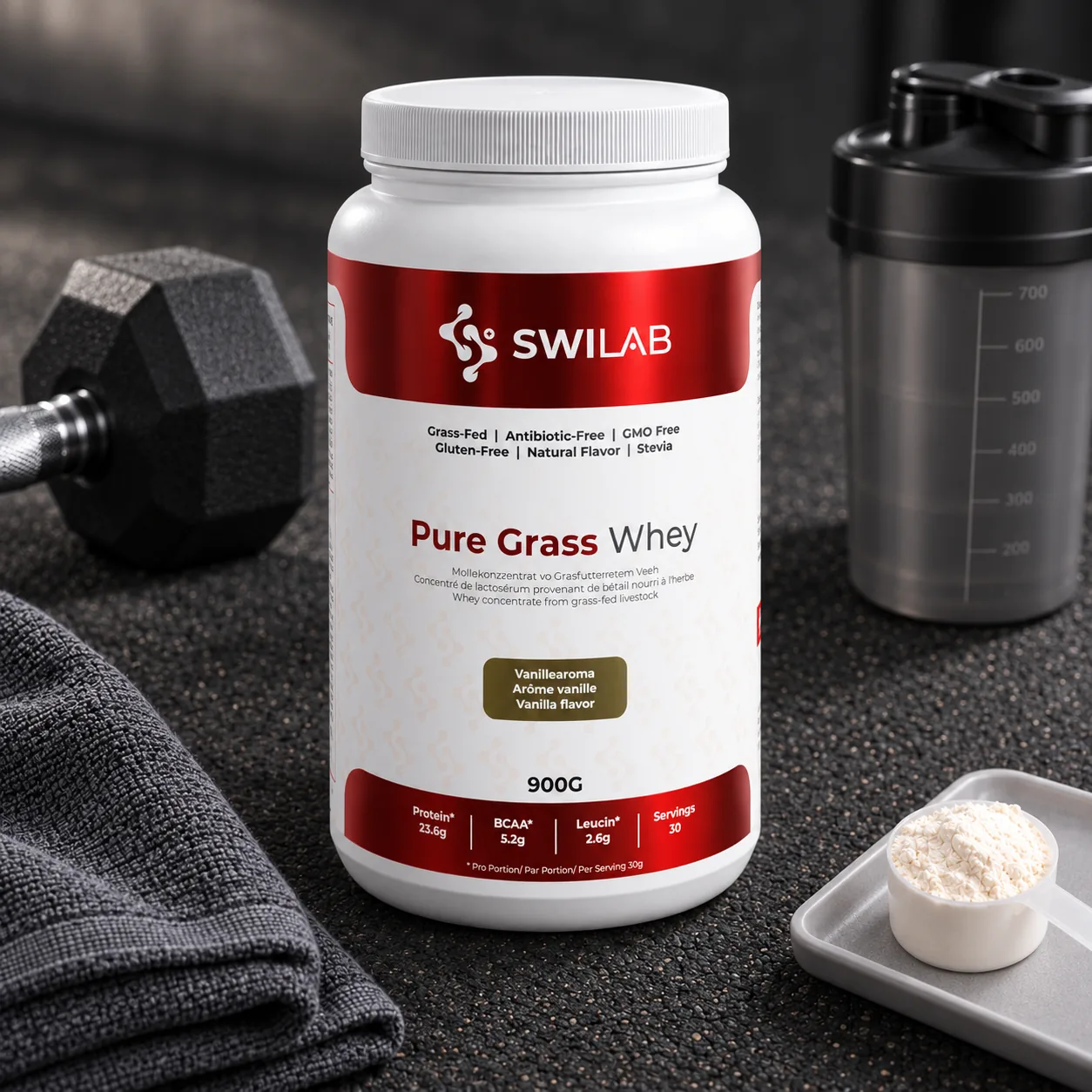 Barattolo di whey protein Pure Grass Whey SwiLab sul pavimento di una palestra, tra un manubrio, un asciugamano, uno shaker e un misurino di polvere.